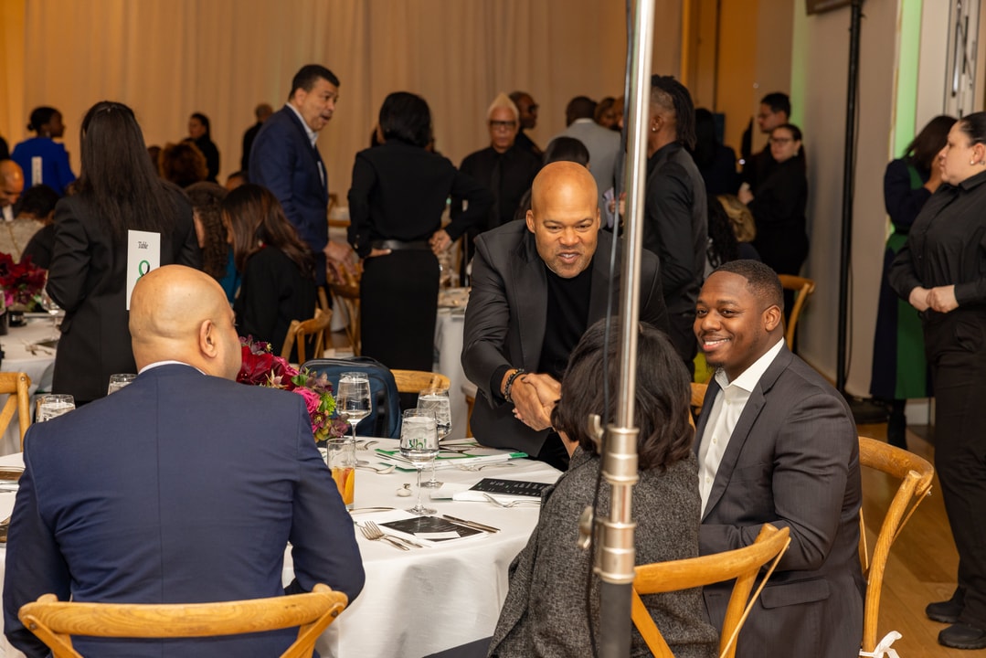 A bald man in a black blazer and black shirt is leaning forward, shaking hands with a seated guest at a round event table. Other guests, including a man in a navy blue suit and another in a gray suit, are engaged in conversation. The background features a busy event hall with attendees, waitstaff, and table decor.