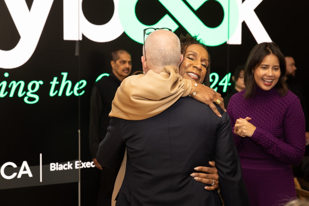 A heartfelt moment as a woman in a beige outfit embraces a bald man in a suit. She is smiling with her eyes closed, conveying warmth and happiness. A digital display in the background shows the words "Black Exec" along with a green-lit event design. Another woman in a purple sweater is seen laughing nearby.