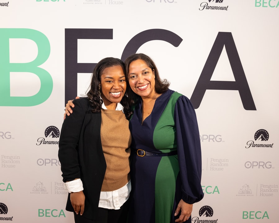 Two women pose together, smiling warmly at the camera in front of a "BECA" event backdrop. One woman is wearing a black blazer over a white shirt with a brown sweater, and the other is dressed in a navy blue and green dress with a belt. They have their arms around each other in a friendly gesture.