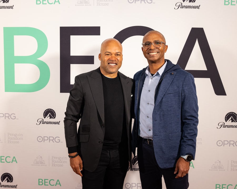 Two men stand side by side, smiling in front of a backdrop with "BECA" and various sponsor logos, including "Paramount" and "Penguin Random House." One man is wearing a black blazer over a black shirt, while the other is dressed in a blue blazer over a light blue button-up shirt.