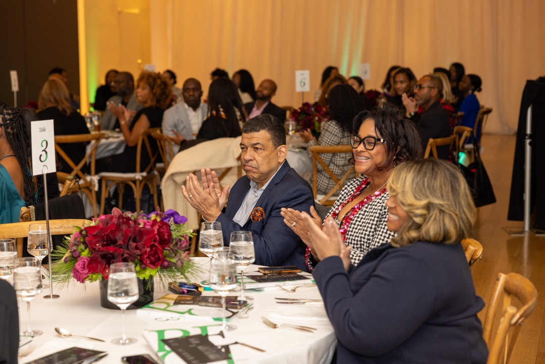 Attendees seated at a round table labeled “3” clap and smile during a formal event, with red floral centerpieces and other guests visible in the background.