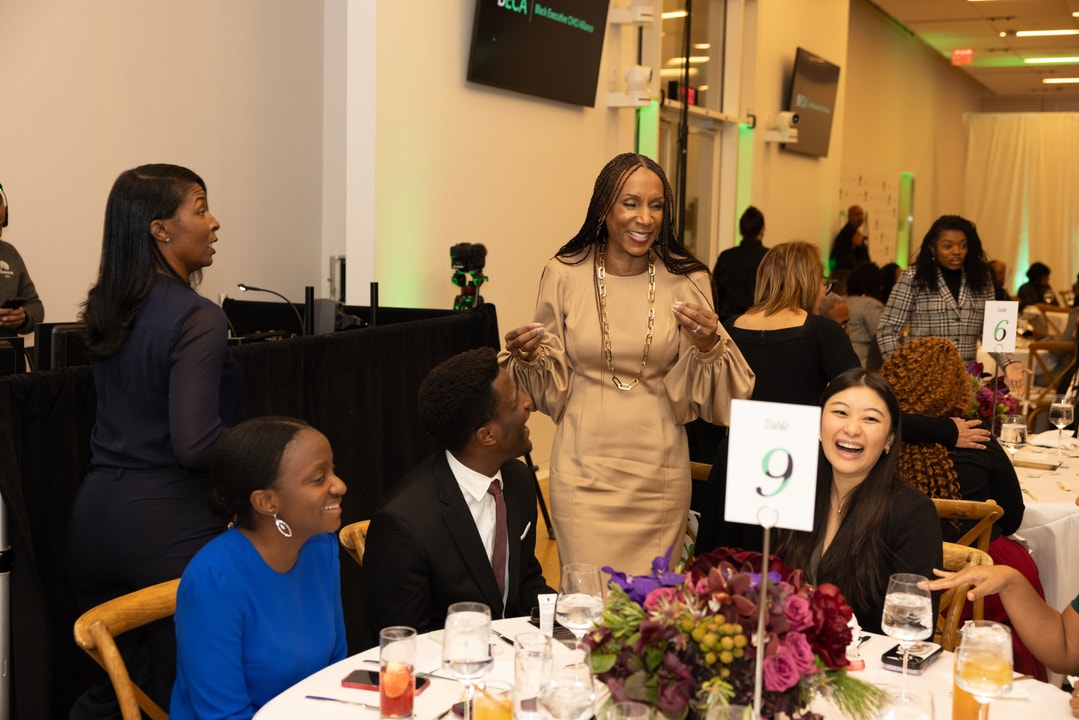 A woman in a beige dress with long sleeves and gold jewelry is standing and engaging with attendees at a round table during a formal event. The guests at the table, dressed in suits and elegant attire, are smiling and laughing. A table sign with the number "9" is visible in the foreground, and other guests and event decor can be seen in the background.