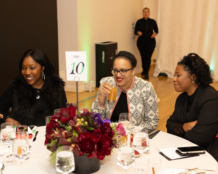 Three women sit at a table engaged in conversation and laughter. The table is decorated with a floral centerpiece, glasses, and event materials. One woman in a black fur vest is smiling, another in a patterned blazer and glasses is sipping a drink, and the third in a black outfit is leaning in attentively. A table sign with the number "10" is visible in the foreground, and a staff member in black attire stands in the background.