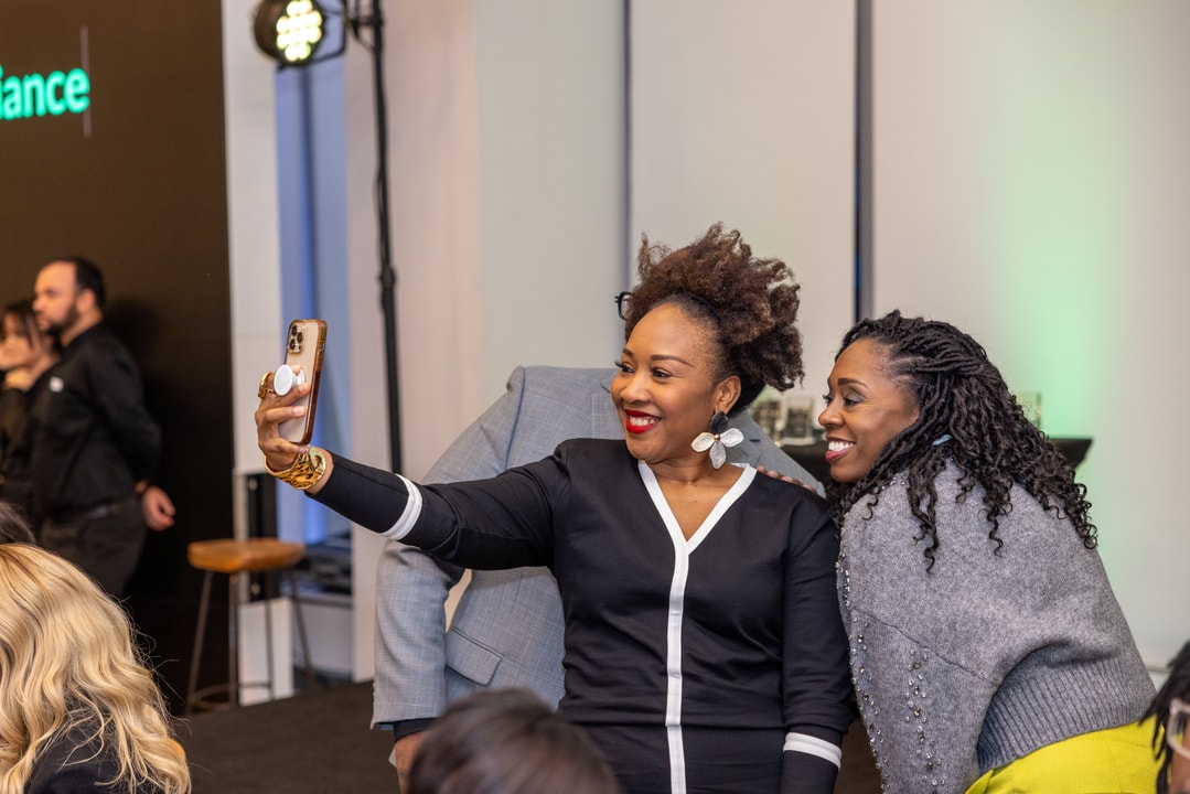 Two women smile brightly as they take a selfie together at an event. One woman in a black and white dress holds a smartphone with a pop socket, while the other, dressed in a gray sweater, leans in. The background features other guests, event lighting, and a stage setup.