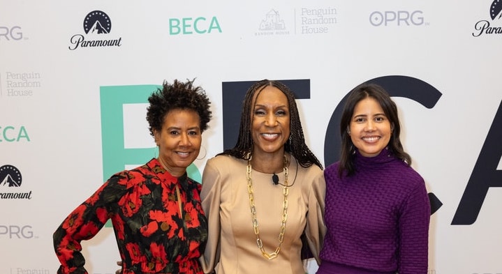 Three women stand smiling together in front of a backdrop featuring various sponsor logos. The woman on the left wears a red floral-print dress, the center woman wears a beige top with a gold necklace, and the woman on the right wears a purple sweater dress.