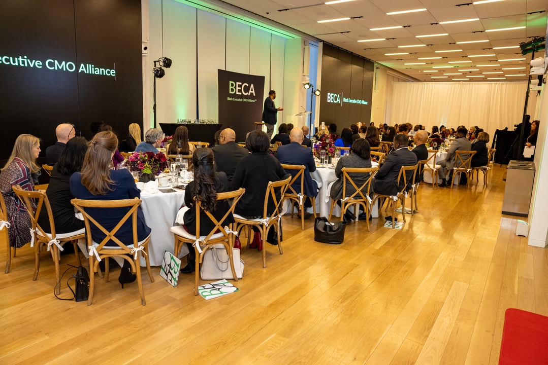 A formal event setup with multiple round tables and guests seated, listening to a speaker on stage. The backdrop on the stage features the "BECA" logo and "Black Executive CMO Alliance." The room has elegant lighting, floral centerpieces on tables, and attendees in business attire.