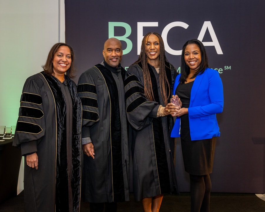 A woman in a red dress and black jacket holds an award while standing beside a woman in academic-style robes at the BECA event.