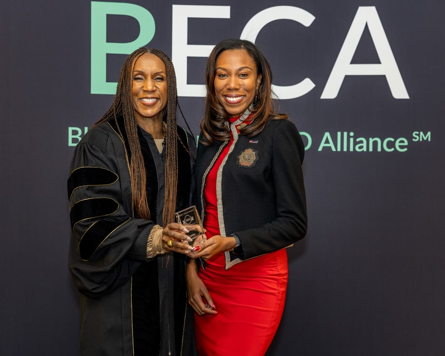 A woman in a red dress and black jacket holds an award while standing beside a woman in academic-style robes at the BECA event.