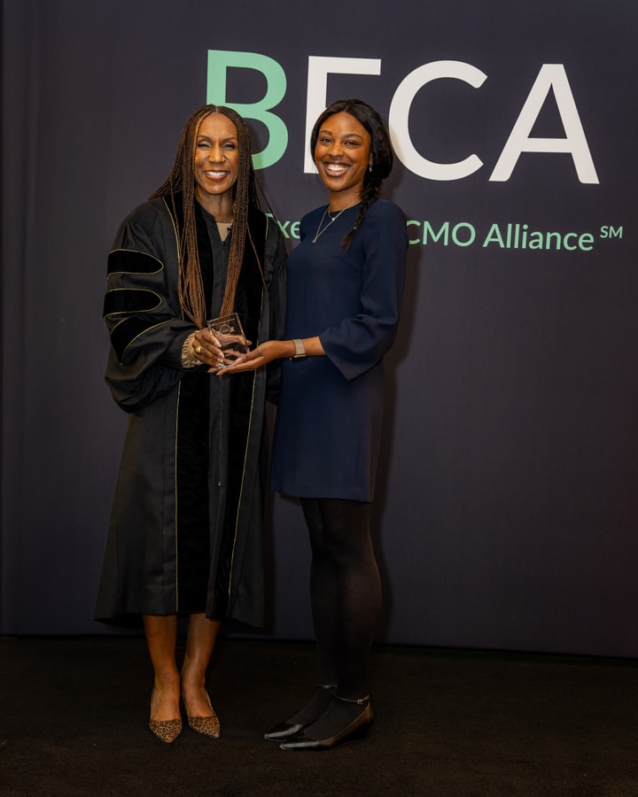 Two women stand together in front of the "BECA" backdrop, smiling. One wears a black academic robe while handing a glass award to the other, who is dressed in a navy blue dress.