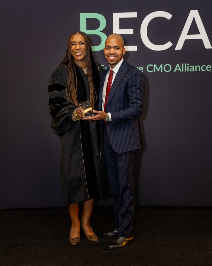 A woman in an academic gown presents an award to a man in a dark suit and red tie, both smiling in front of a "BECA" backdrop.