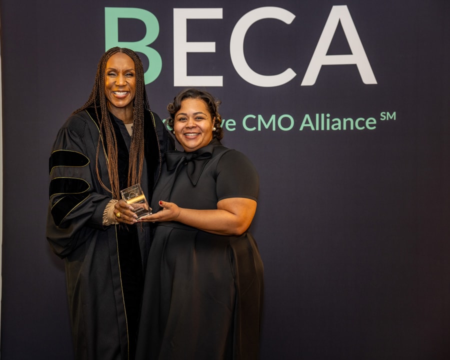 A woman in a black dress smiles while holding an award alongside a woman in academic-style robes in front of the "BECA" backdrop.