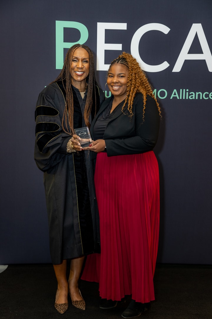 Two women stand together, smiling at the camera. One woman, dressed in a black academic gown, holds an award while the other, wearing a black blazer and a red skirt, stands beside her.