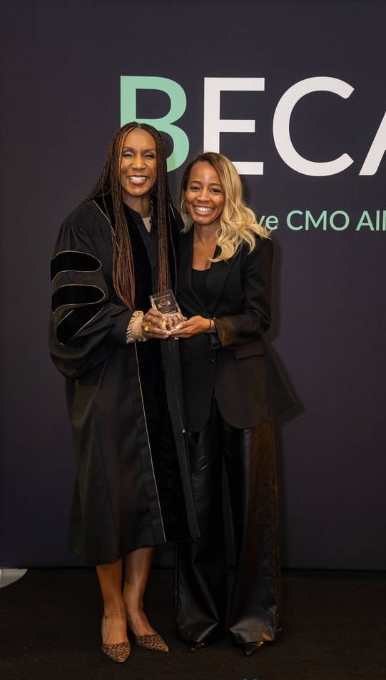 Two women, one in an academic robe and the other in a black suit, pose together holding an award in front of a "BECA" backdrop.