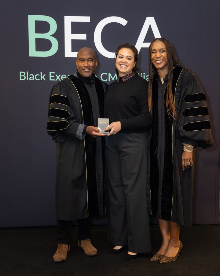 A man and two women stand in front of a "BECA Black Executive CMO Alliance" backdrop. The man and one of the women wear black ceremonial robes with gold trim. The woman in the center, dressed in a black sweater and gray pants, holds an award and smiles.