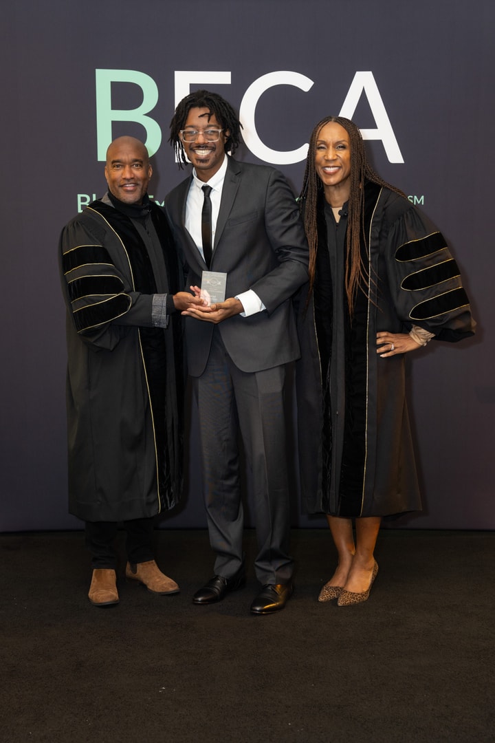 Three individuals pose in front of a "BECA Black Executive CMO Alliance" backdrop. The person in the center, a young man in a gray suit and black tie, holds an award. The two individuals beside him, a man and a woman, are dressed in black ceremonial robes with gold trim and smiling.