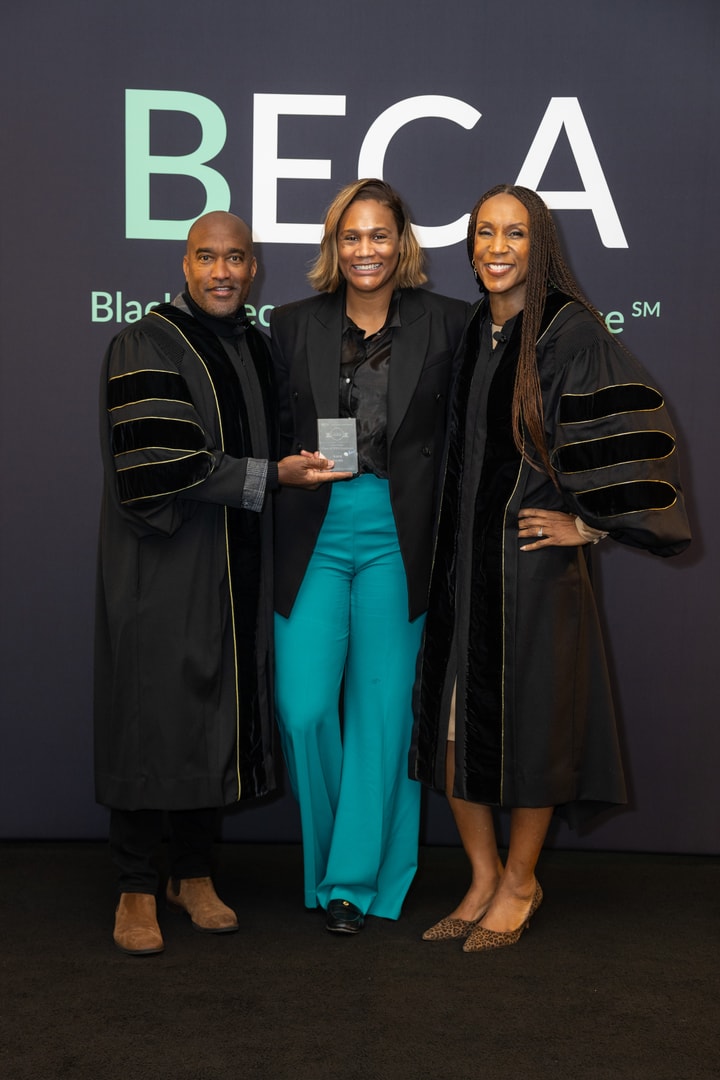 Three people stand together in front of a backdrop with "BECA Black Executive CMO Alliance" written on it. The person in the center, a woman in a black blazer and turquoise pants, holds a small award. The two individuals beside her, a man and a woman, are wearing black ceremonial robes with gold accents and smiling.