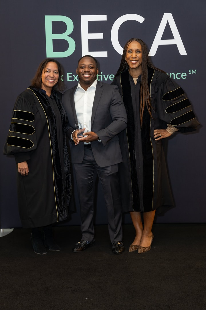 A man in a suit holds an award while standing between two women in black academic gowns. All three are smiling in front of the BECA backdrop.