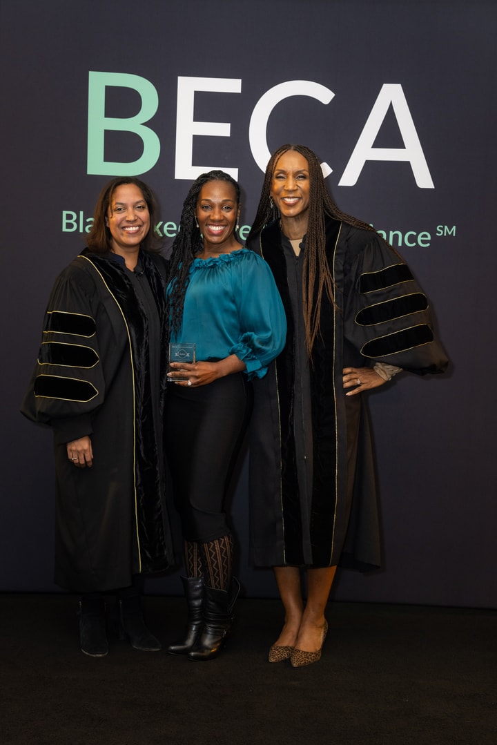 Three women stand together in front of a "BECA" backdrop, smiling. One holds a glass award while the other two wear black academic robes.