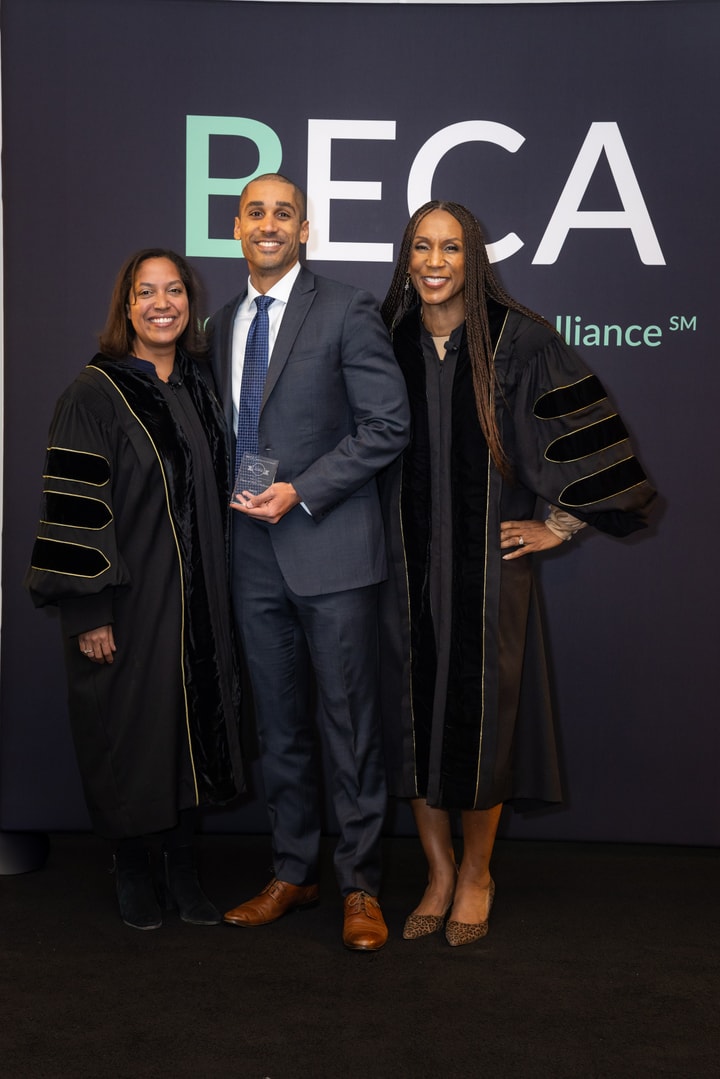 A man in a dark blue suit holding an award stands between two women in black academic robes, all smiling in front of a "BECA" backdrop.
