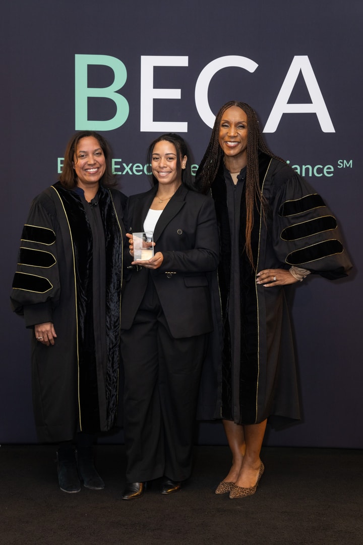 Three women pose with an award in front of a "BECA" backdrop, two in academic gowns and one in a black suit holding the award.