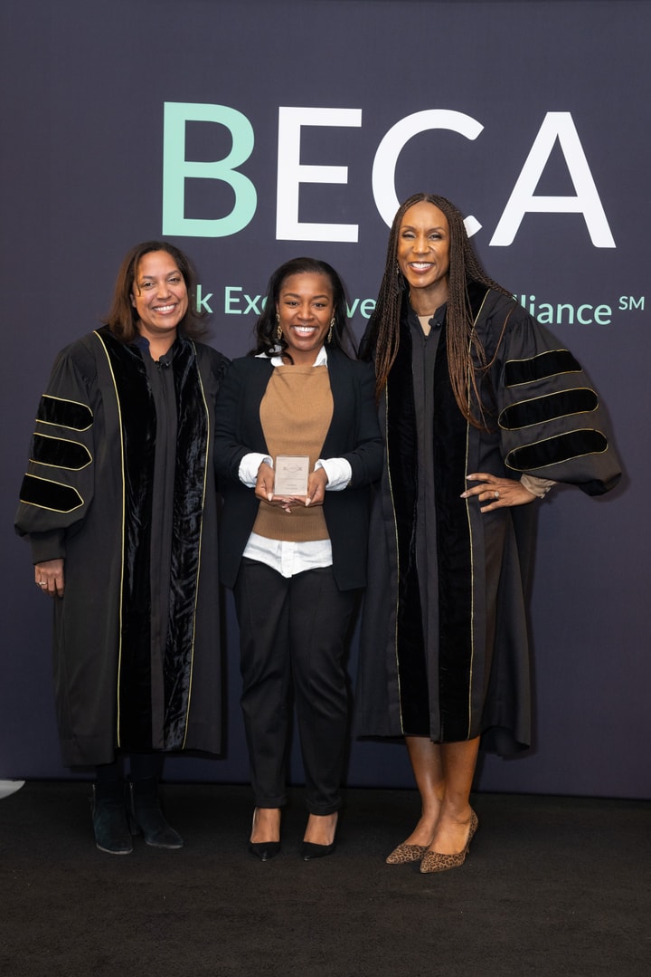A woman in a black academic gown presents an award to another woman dressed in a beige sweater and black pants. They both smile in front of the BECA backdrop.