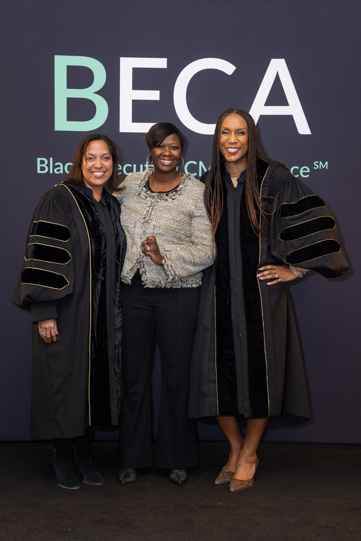 Three women stand together in front of a "BECA" backdrop, two wearing academic gowns and one in a tweed jacket, all smiling.