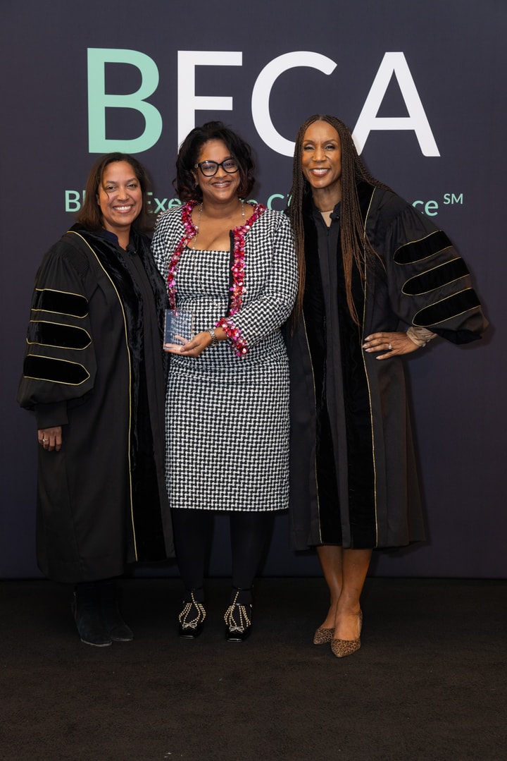 Three women pose in front of the "BECA" backdrop. The central woman, wearing a black and white houndstooth dress, holds an award while the two on either side wear black academic robes.