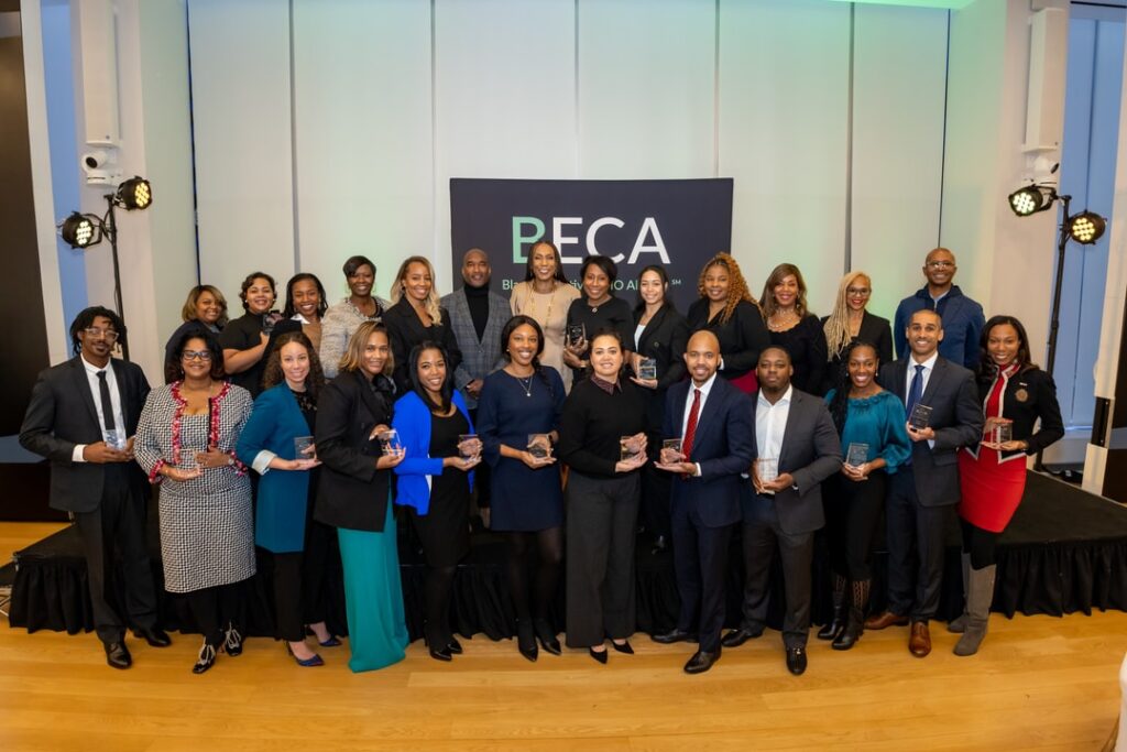 A group of professionals pose for a photo, each holding a glass award. The backdrop features a sign with the text "BECA" and "Black Executive CMO Alliance." The attendees are dressed in formal business attire, with many smiling and looking at the camera. The setting is an event space with a stage, spotlights, and a modern, well-lit interior.