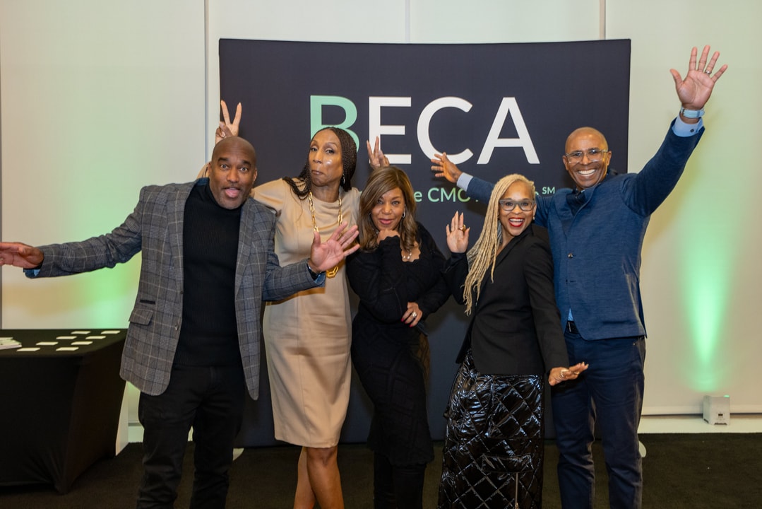 Five people pose playfully in front of a "BECA" event backdrop, making gestures like raising hands, peace signs, and bunny ears. They are dressed in stylish professional attire, with some wearing blazers and dresses. The group appears joyful, engaging in a fun and energetic moment at the event.