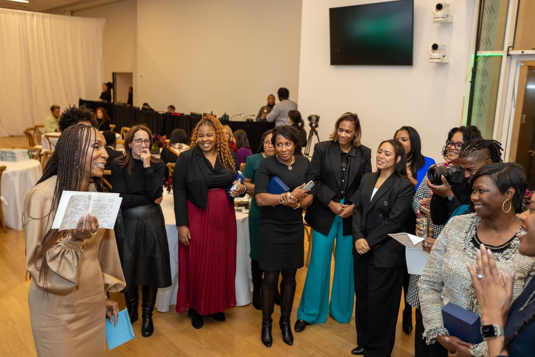 A woman in a beige dress with long sleeves and gold jewelry is addressing a group of attendees at a formal event. She holds a notebook and gestures while speaking, engaging the diverse group of women in front of her, who are dressed in a mix of professional and stylish attire. Some attendees are smiling, while others hold notebooks, papers, or cameras. The background includes event tables, a large screen, and photographers capturing the moment.