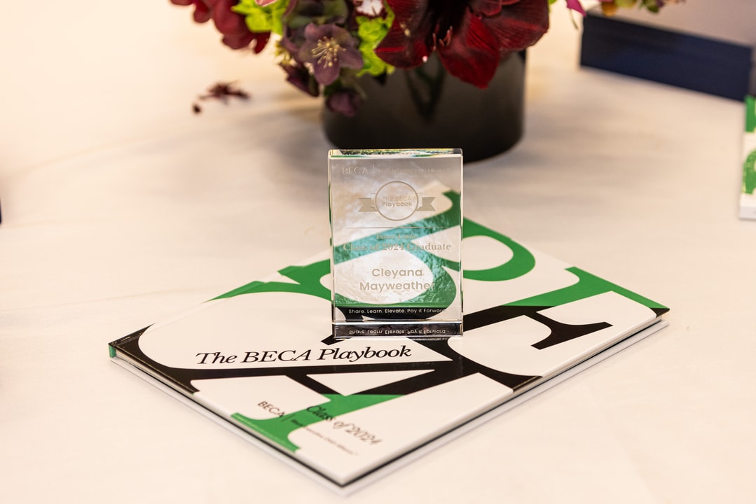 A clear glass award engraved with the name "Cleyana Mayweather" sits atop a book titled The BECA Playbook. The award acknowledges a graduate from the BECA program, with the words "Share, Learn, Elevate, Pay It Forward" inscribed. A bouquet of deep red and purple flowers decorates the table in the background.