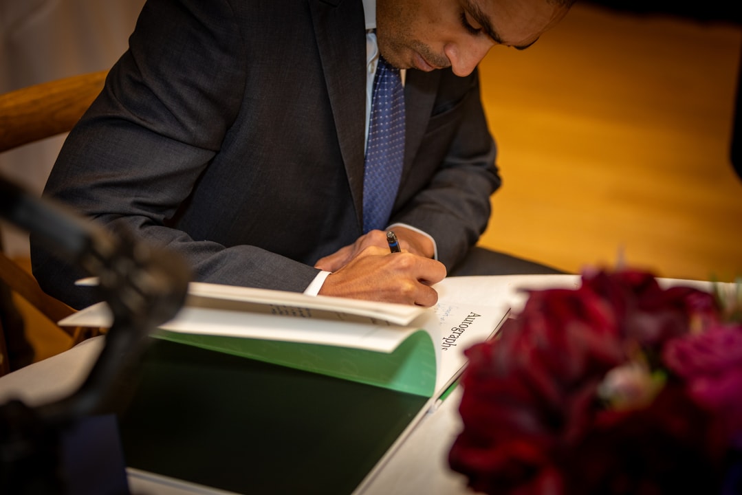 A man in a dark suit and blue tie is signing a book with a focused expression. His hands are holding a pen as he writes on an open page. The setting is a formal event, with a table covered in a white cloth and a blurred bouquet of red flowers in the foreground.