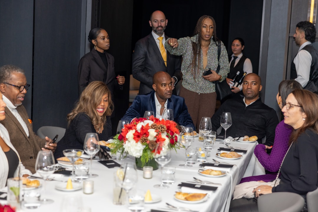 A woman in a shimmering outfit gestures while speaking at a formal dinner gathering. Guests at the table listen, while others stand in the background.