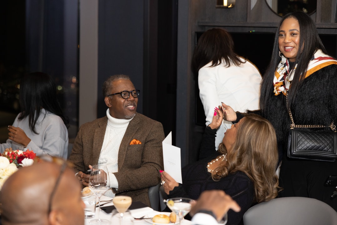 A man in a brown blazer and glasses smiles while speaking with a woman at a formal dinner. Another woman stands nearby, greeting guests with a smile.