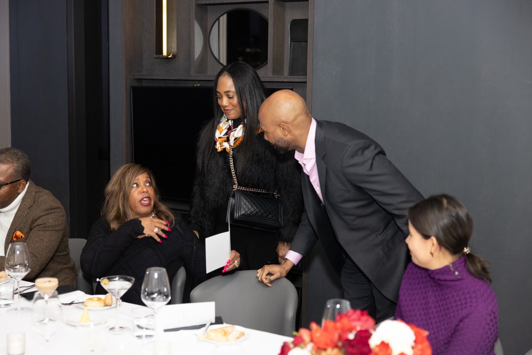A man in a suit leans in to greet a seated woman at an elegant dinner event, while another guest stands nearby smiling. The table is set with fine dining arrangements and red floral centerpieces.