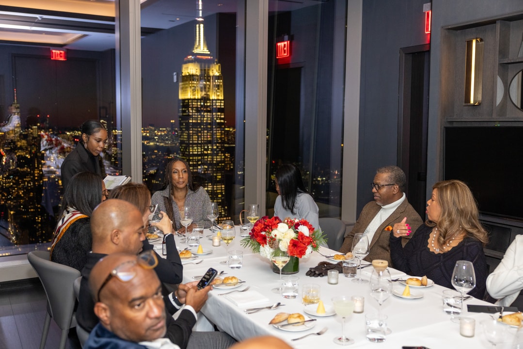A group of elegantly dressed guests converses around a beautifully set dinner table, with the illuminated Empire State Building visible through the window.