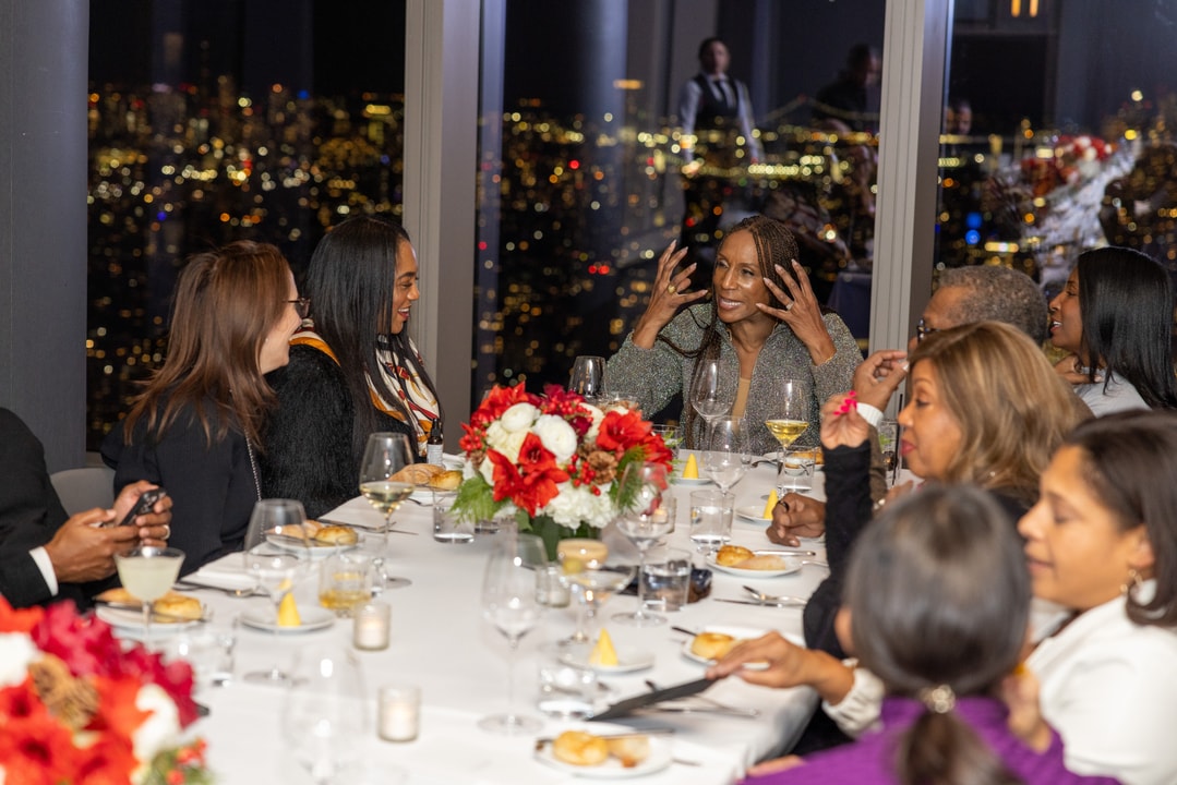 A group of people enjoys a dinner gathering with a stunning nighttime cityscape in the background. A woman in a shimmering outfit gestures expressively while speaking.