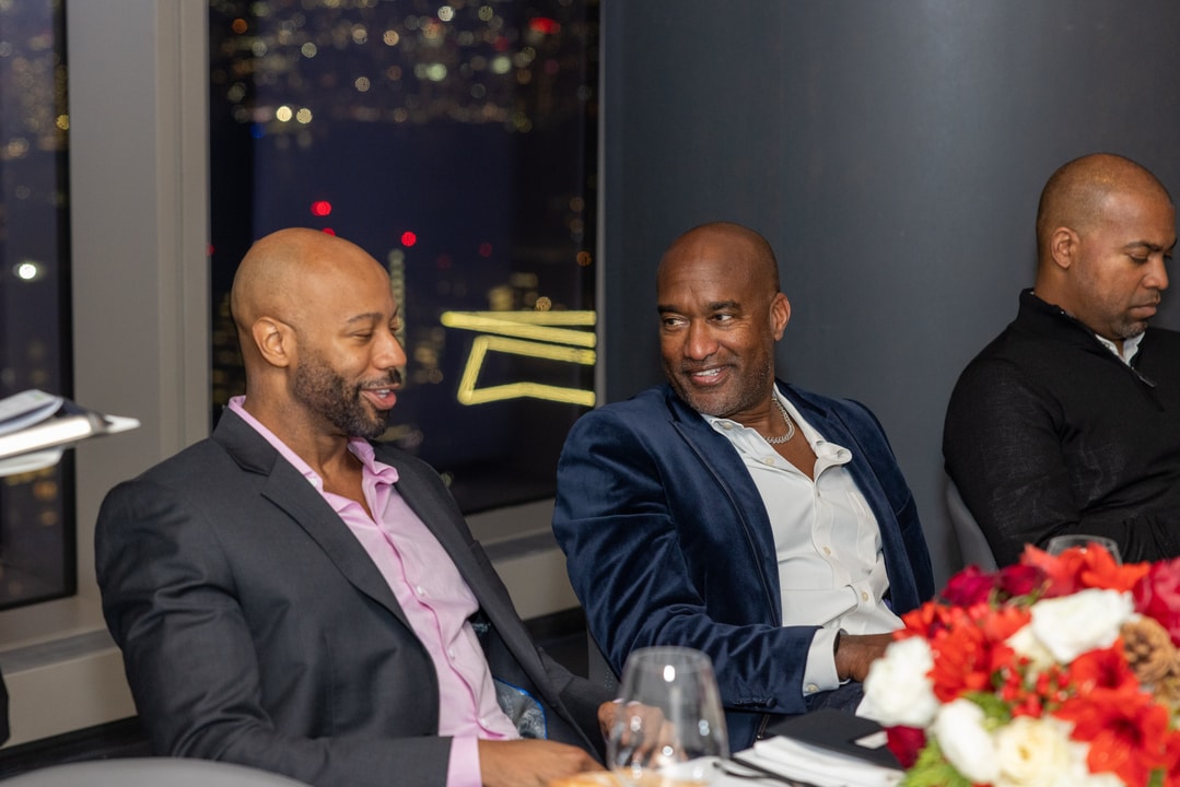 Two men in suits engage in a friendly conversation at an upscale dinner event. A nighttime city skyline with illuminated buildings is visible through the window behind them.