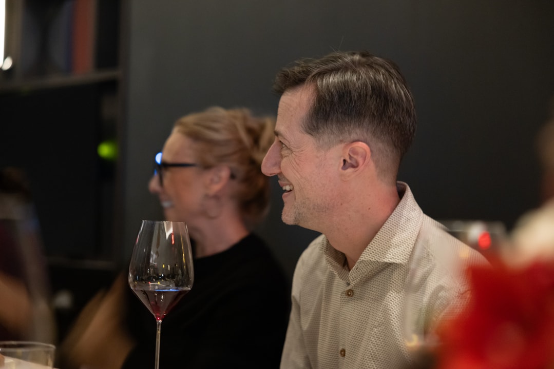 A man smiles while engaged in conversation at a formal dinner, with a glass of red wine in the foreground. A woman seated next to him laughs, enjoying the moment.