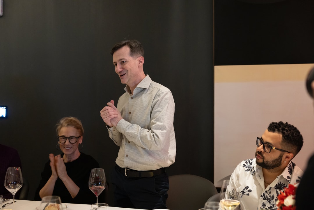 A man stands and claps while giving a speech at a formal dinner, as guests seated at the table smile and applaud.