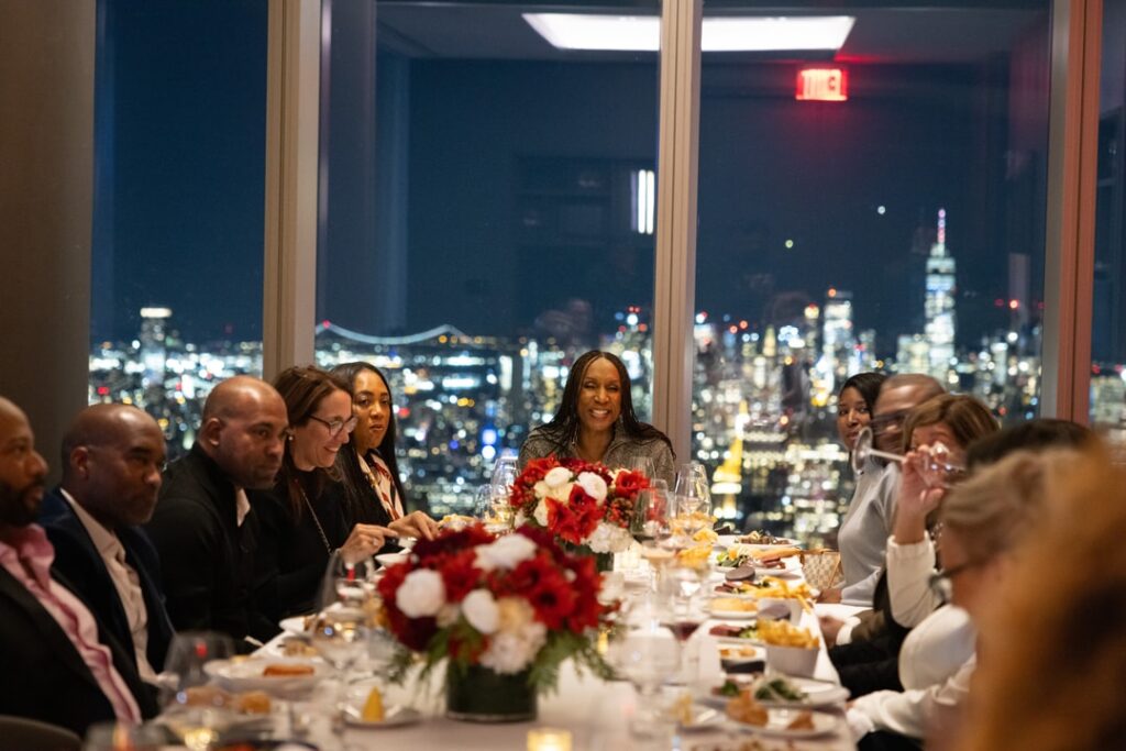 Elegant Dinner with City Skyline View – A group of people enjoys a formal dinner at a beautifully set table with red and white floral centerpieces. A woman in the center smiles, and the backdrop features a stunning nighttime city skyline with illuminated buildings and bridges.