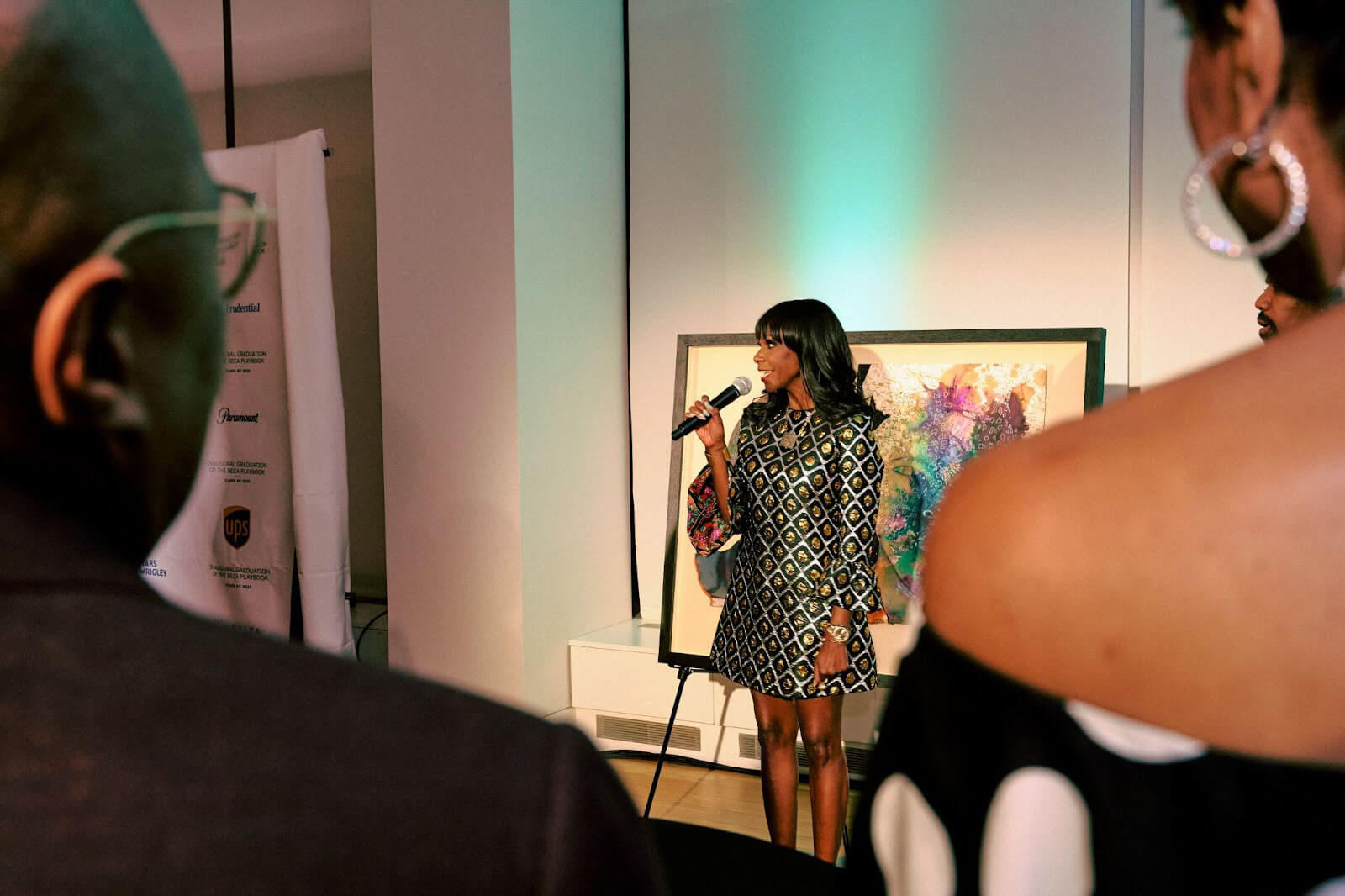 A woman in a patterned dress speaking into a microphone in front of a vibrant piece of artwork, with the audience partially visible in the foreground.