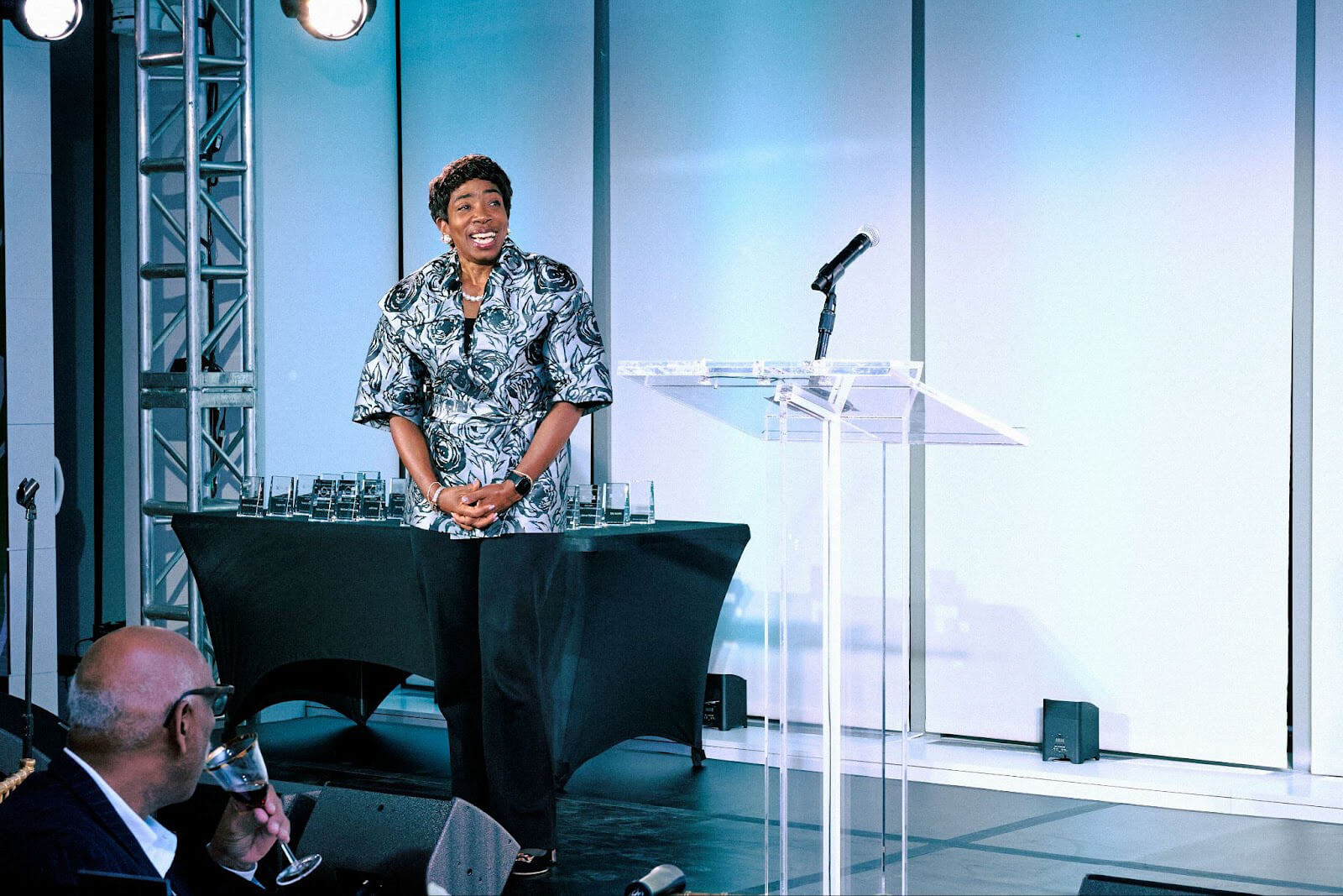 A speaker on stage wearing a floral-patterned outfit, smiling as they address the audience, with awards displayed on a nearby table.