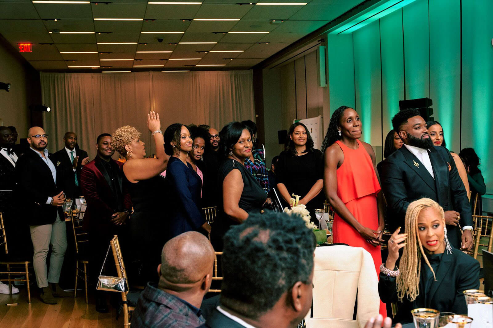 A group of elegantly dressed attendees standing and smiling during a formal gathering, with vibrant lighting in the background.