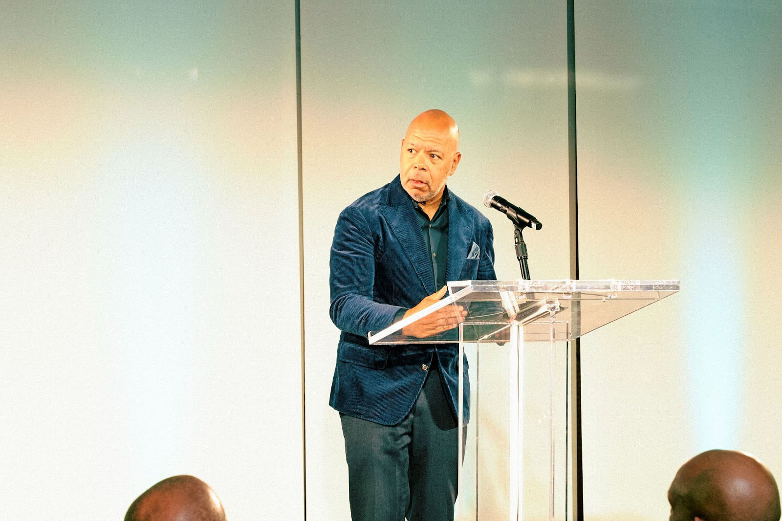 A speaker in a blue blazer addressing the audience at a clear podium in a brightly lit venue.