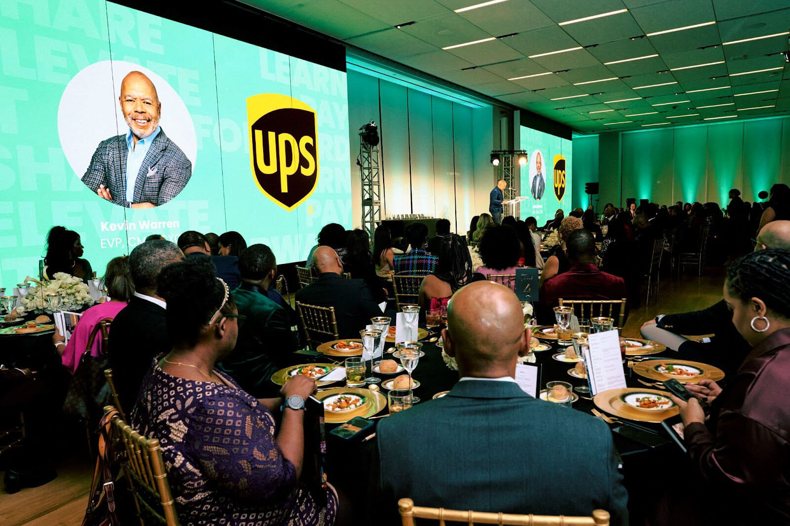 A large audience seated at decorated tables, focusing on a speaker at a stage featuring a UPS logo on a green screen backdrop.