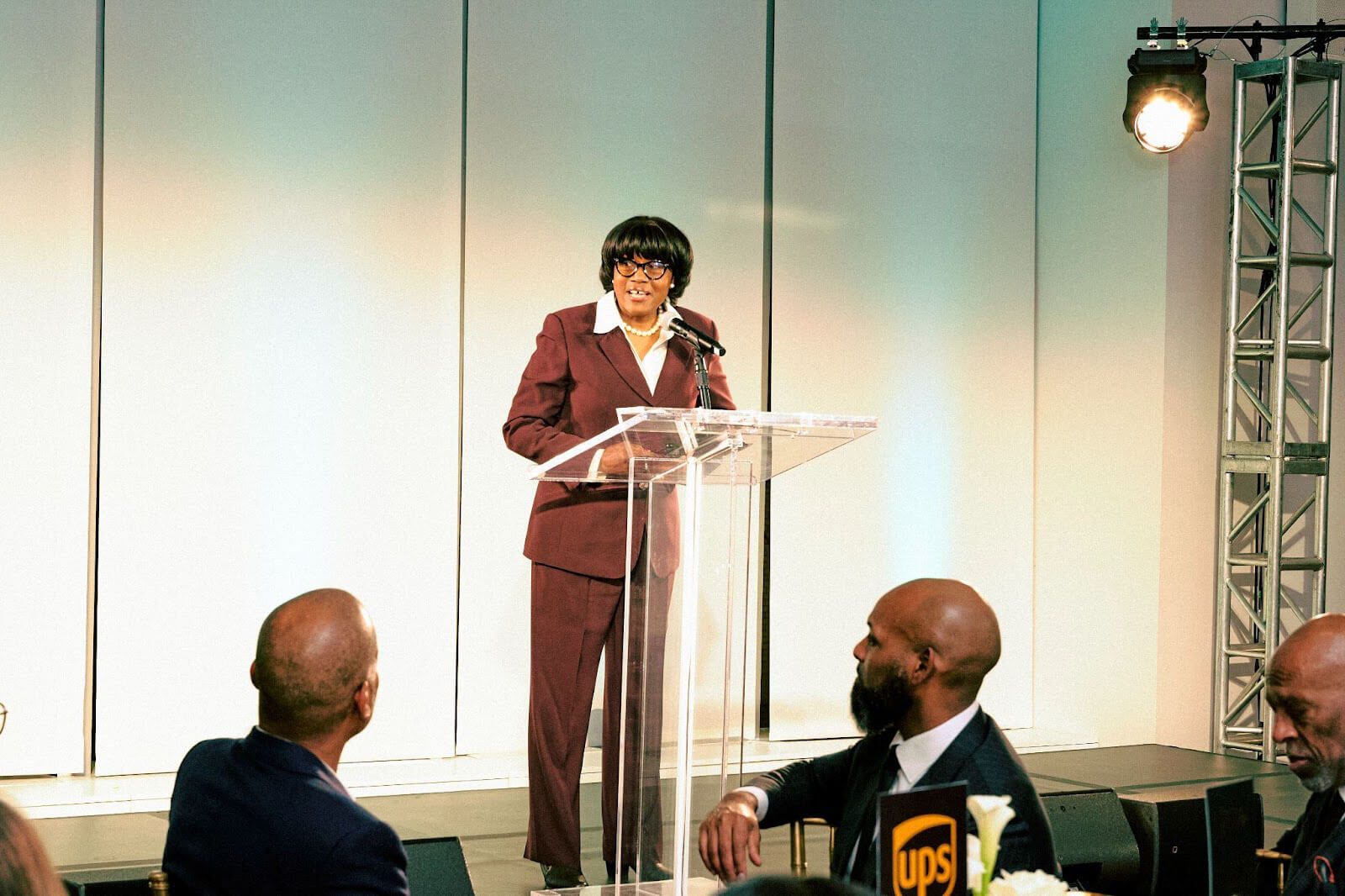 A woman in a burgundy suit speaking at a clear podium during an event, with a spotlight illuminating the stage.