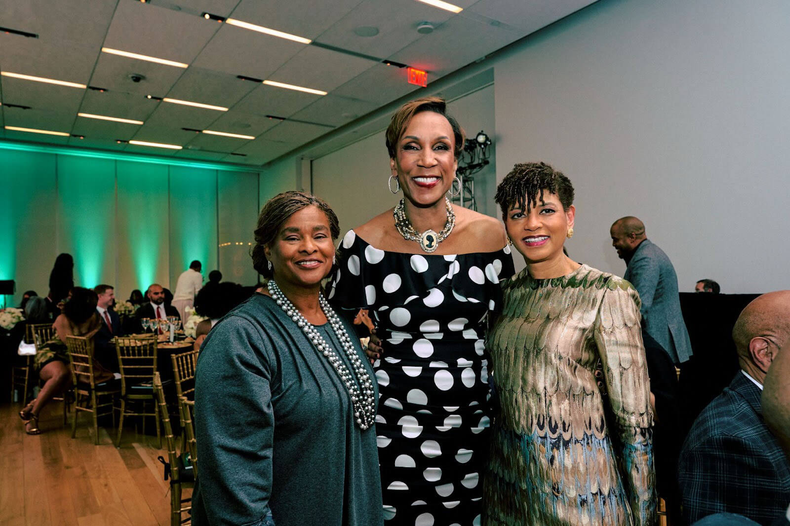 Three women posing together, dressed in formal attire and smiling, against the backdrop of an event space with soft green lighting.