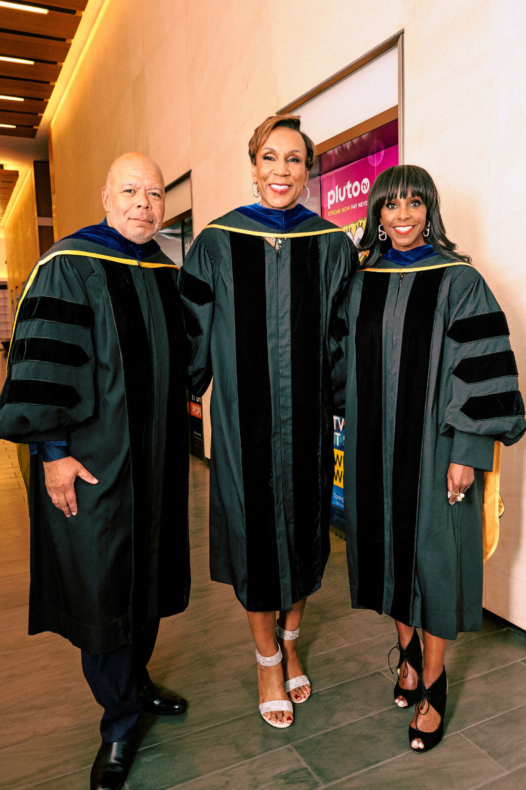 Three individuals in academic regalia posing together, smiling, and standing in a well-lit hallway.