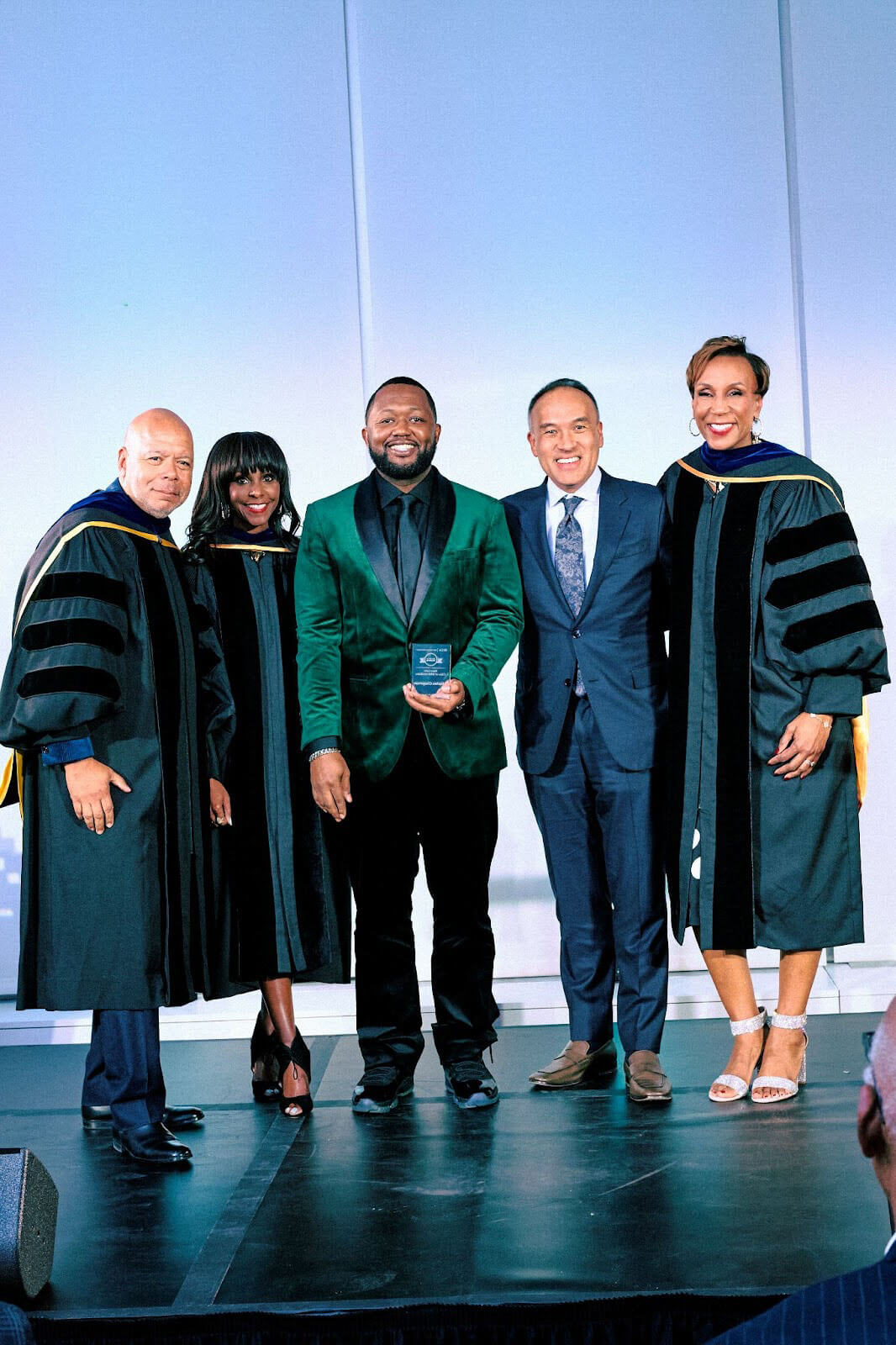 A group photo of five individuals, with three wearing academic regalia and one holding an award, standing on a stage.